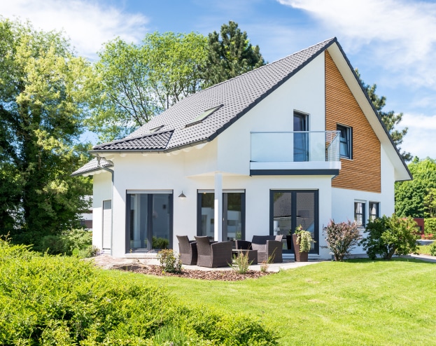 Belle maison moderne blanche à deux étages avec toit sombre, bardage bois clair et balcon en verre, entourée de verdure.