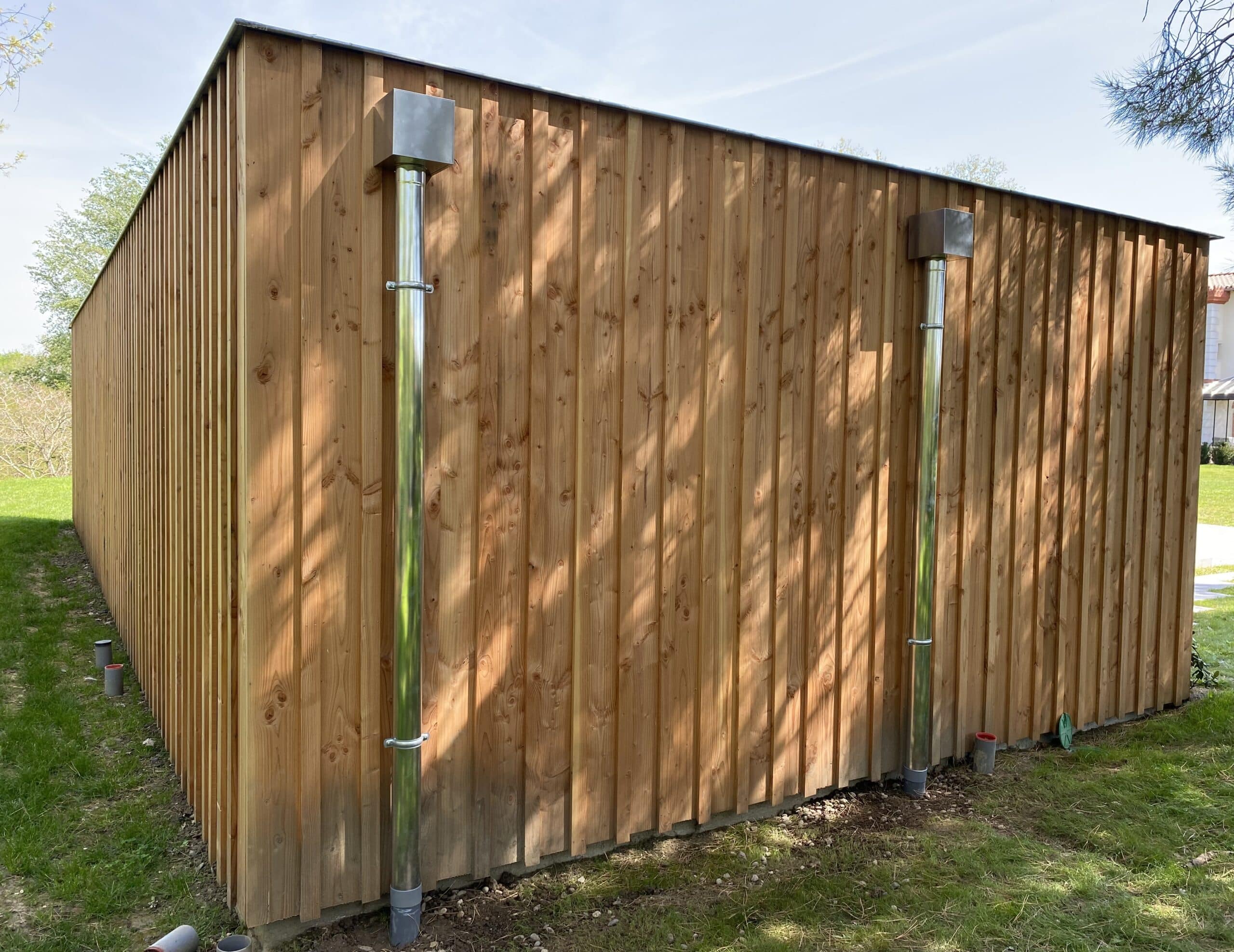 Structure en bois clair avec bardage vertical, deux tuyaux d'évacuation métalliques et caps de tuyaux gris dans l'herbe.