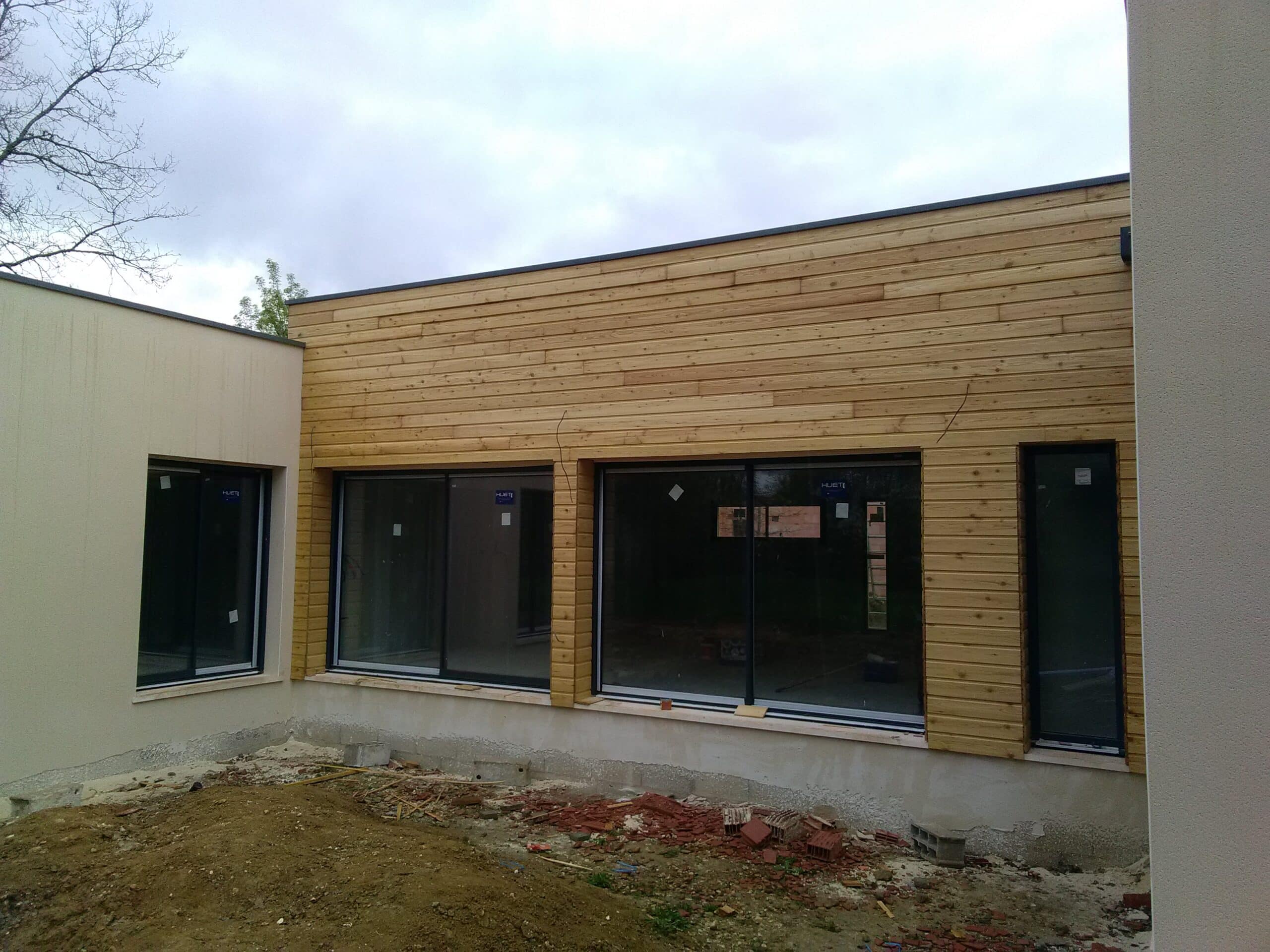 Façade d'une maison moderne en construction avec bardage bois clair et enduit blanc, fenêtres noires (mention HUET visibles).