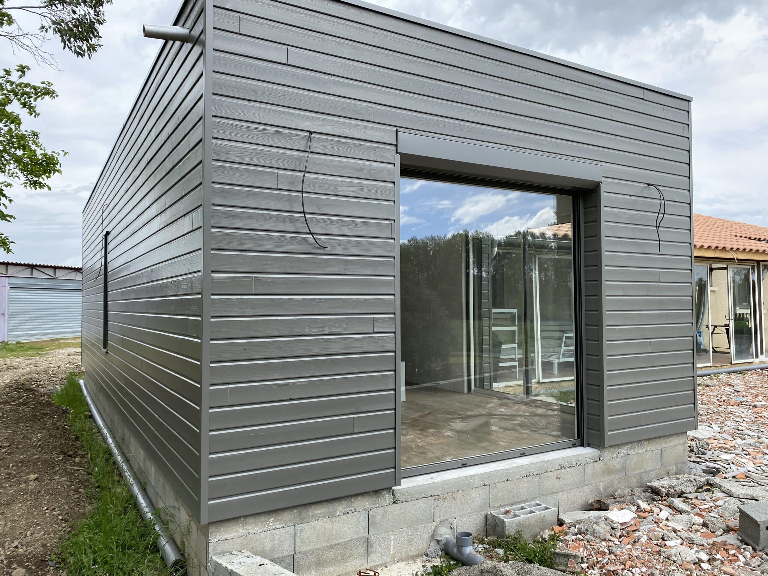 Maison modulaire grise en construction, bardage bois, grande baie vitrée et fondations en parpaings, chantier visible.