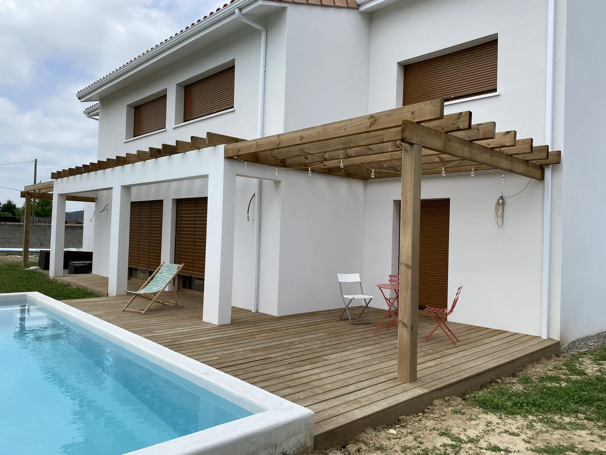 Maison blanche moderne avec terrasse bois, pergola, et piscine extérieure en construction.