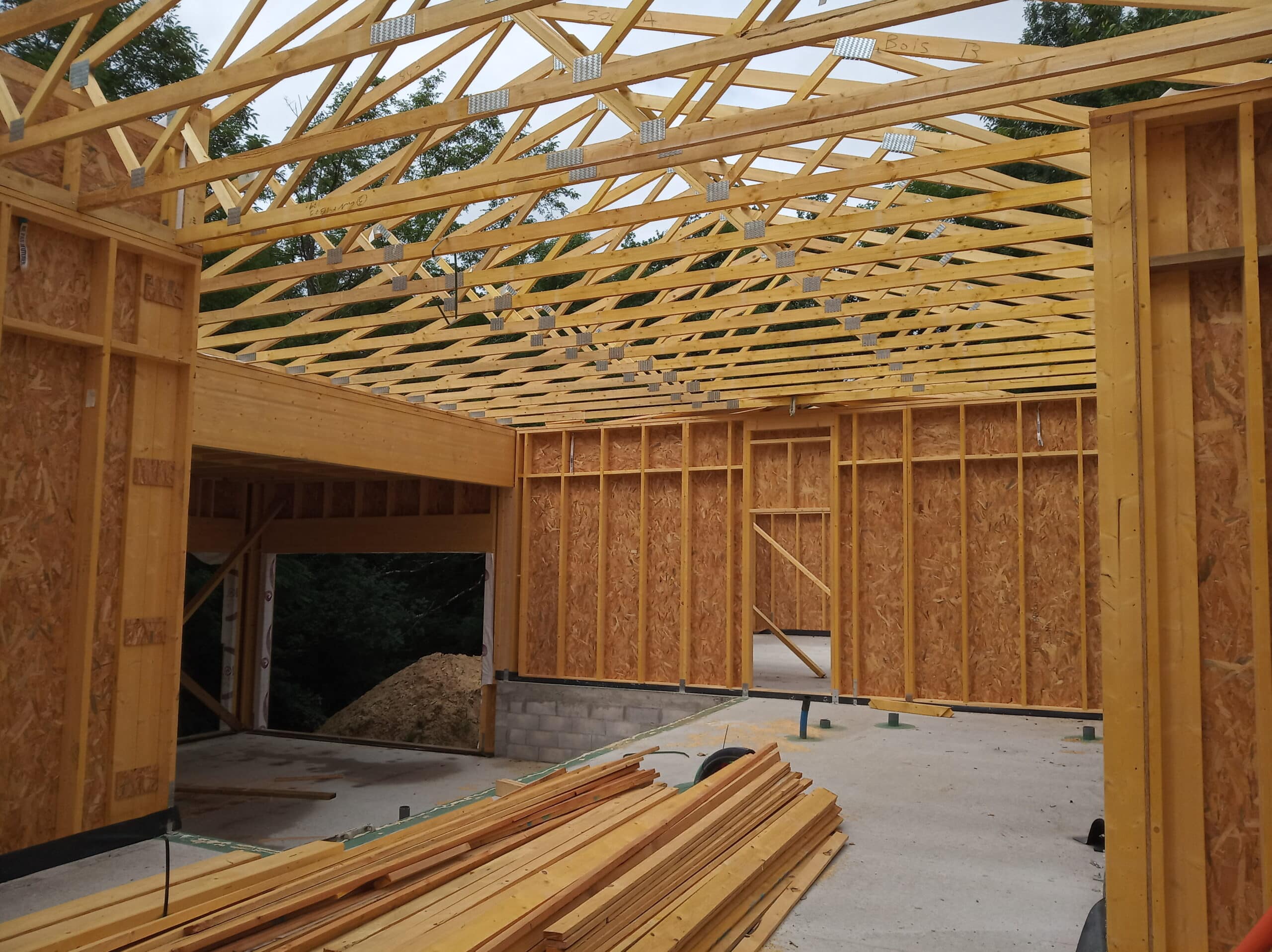 Construction d'une ossature bois avec charpente en fermes métalliques et parpaings, bois de charpente empilé au sol. Texte visible: "Bois".