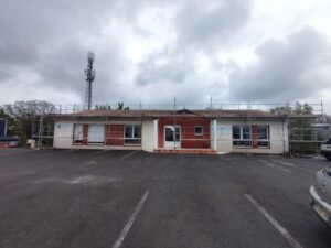 Bâtiment ABM en rénovation avec échafaudage sur parking sous un ciel nuageux, antenne relais en arrière-plan.