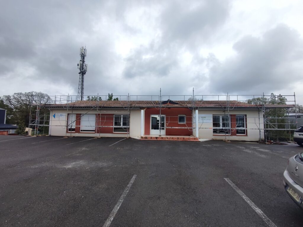 Bâtiment ABM en rénovation avec échafaudage sur parking sous un ciel nuageux, antenne relais en arrière-plan.