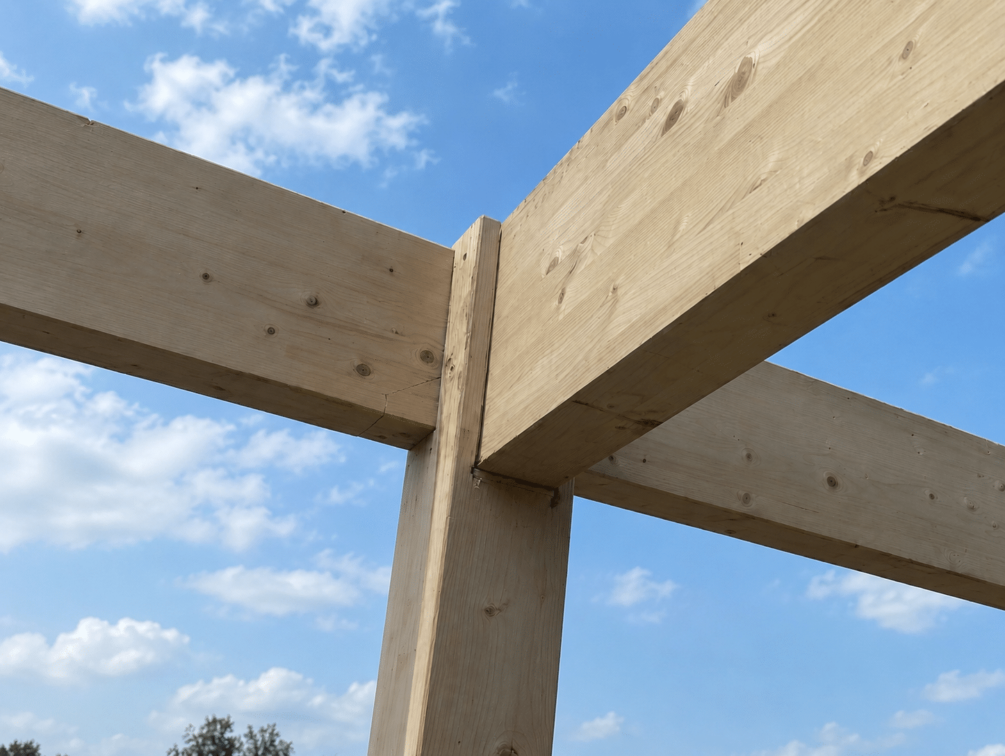 Assemblage de poutres en bois clair d'une charpente sous un ciel bleu nuageux.