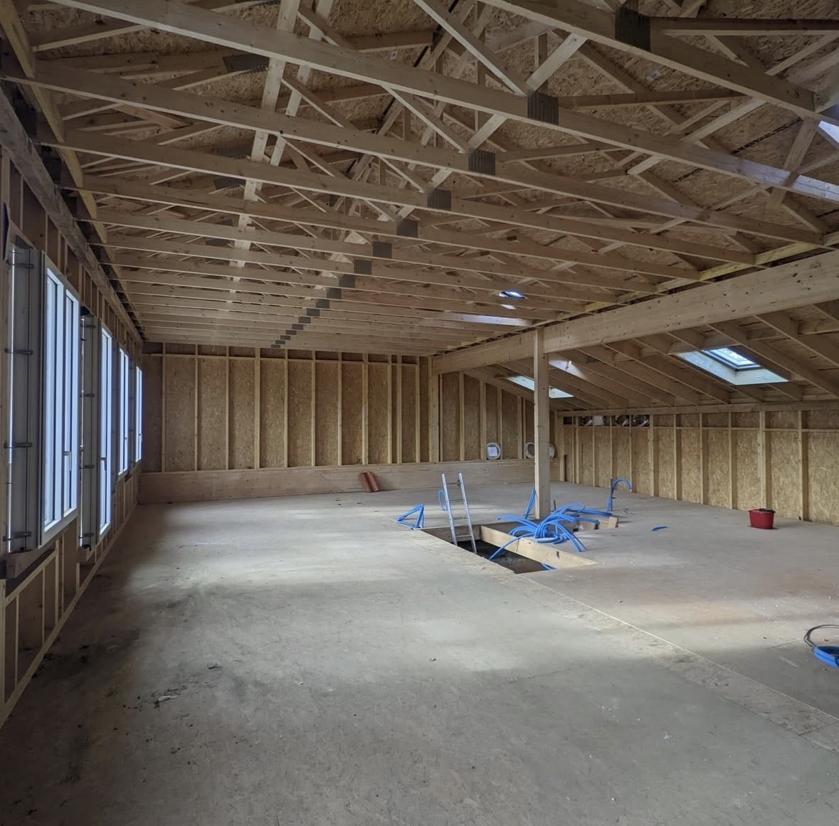 Intérieur de maison bois en chantier avec charpente apparente, murs OSB et gaines bleues au sol.