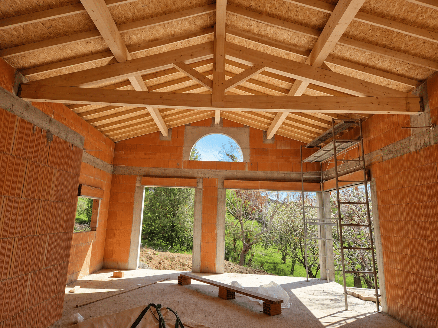 Maison en chantier avec charpente bois, murs en briques et vue sur jardin verdoyant.