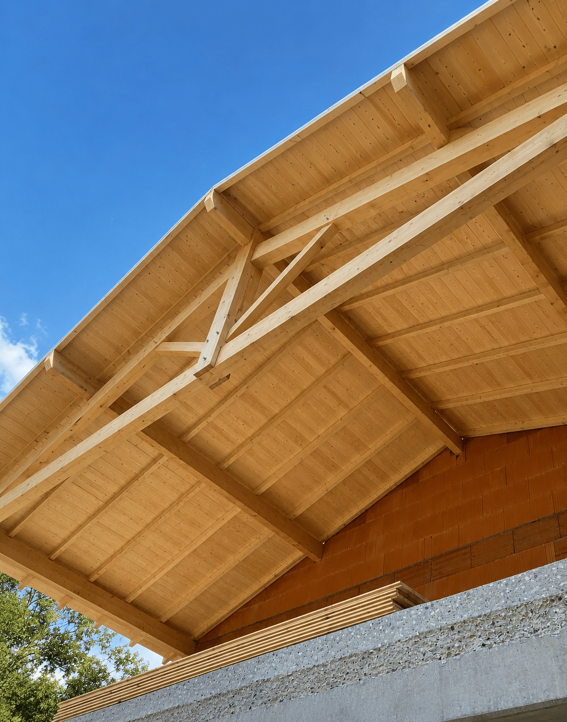 Charpente en bois clair avec poutres apparentes sous un ciel bleu, surmontant un mur de briques en construction.