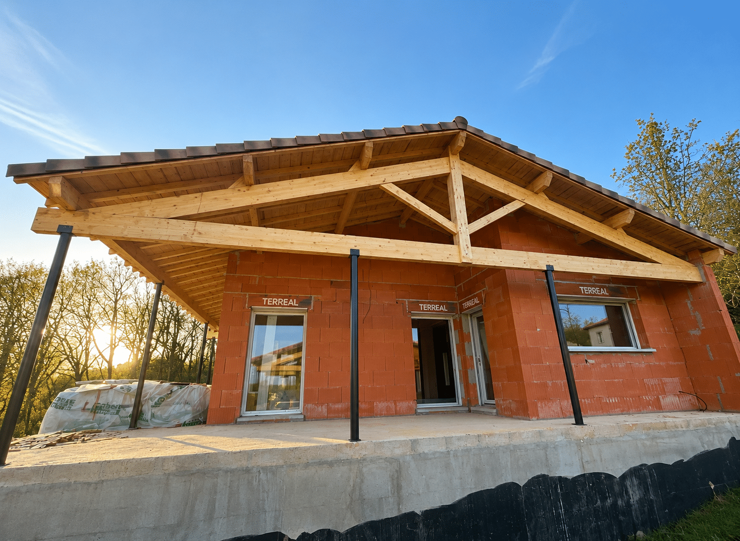 Maison en briques Terreal, charpente bois apparente et piliers noirs sur fondation béton.