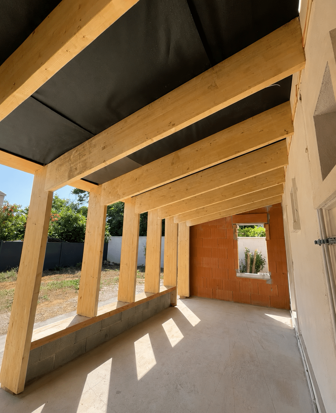 Charpente bois de terrasse, poutres massives et mur de briques en construction sous un soleil éclatant.