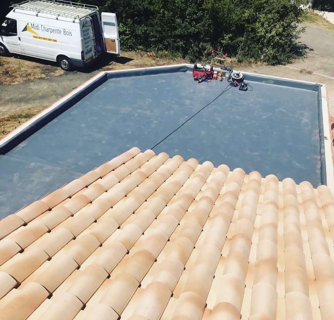 Toit plat en membrane grise, camionnette Midi Charpente Bois et tuiles.