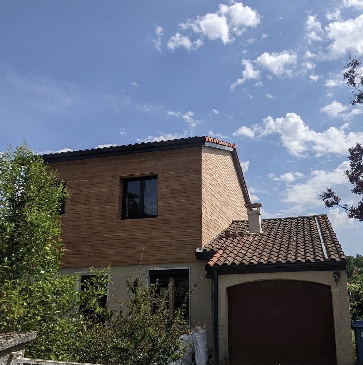 Maison à étage avec bardage bois clair, toit de garage en tuiles et ciel bleu parsemé de nuages.