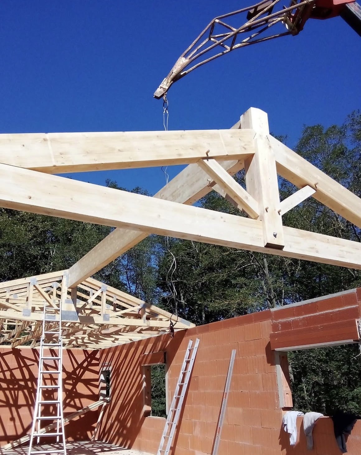 Grue levant une charpente bois sur maison en brique rouge.
