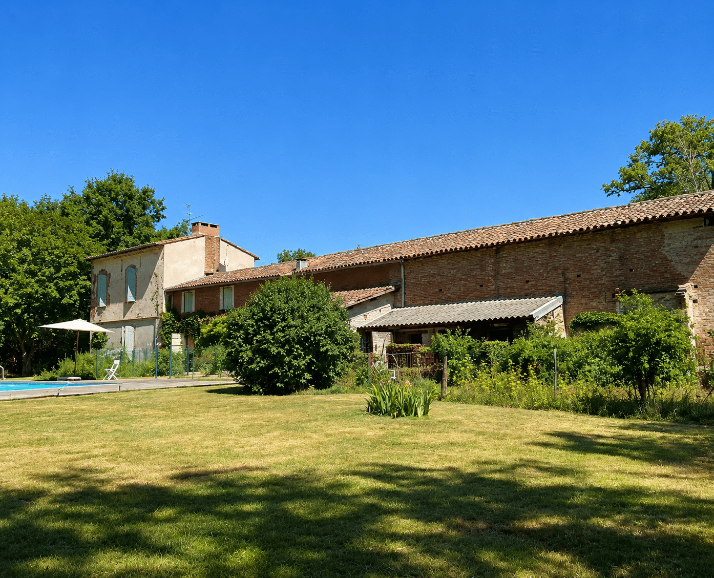 Ferme traditionnelle en briques avec piscine et grand jardin sous un ciel bleu dégagé.