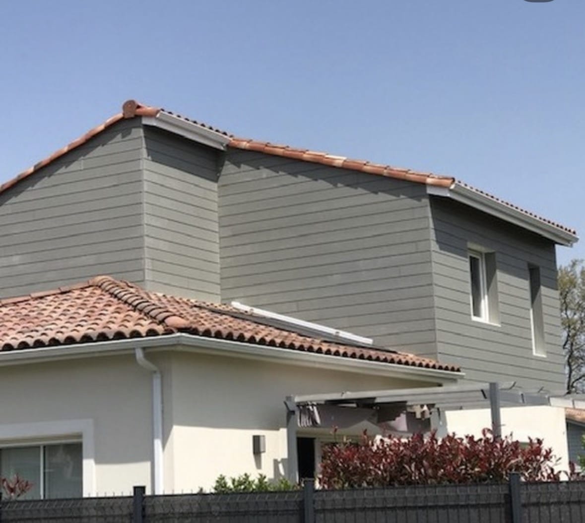 Maison à étage, bardage gris, toit en tuiles rouges et façade claire sous ciel bleu.
