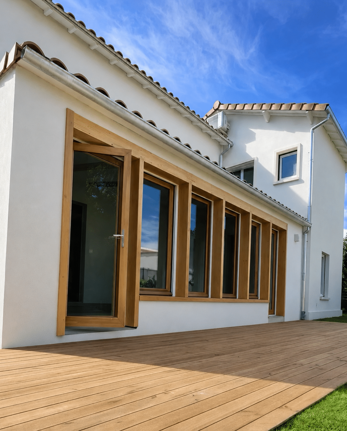 Maison moderne blanche avec terrasse et fenêtres en bois clair sous un ciel bleu.