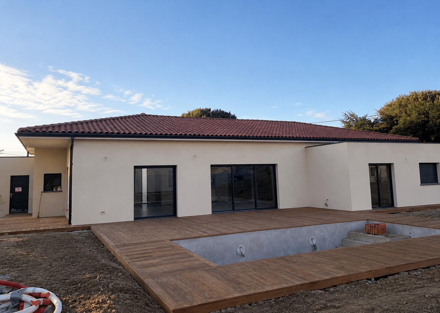 Maison moderne beige, toit en tuiles rouges, grande terrasse en bois et piscine vide sous un ciel bleu.