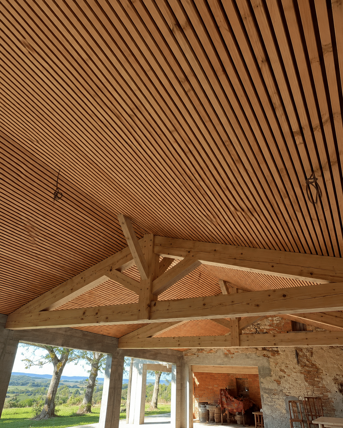 Plafond à lattes de bois et charpente apparente dans une structure ouverte avec vue sur la campagne vallonnée.