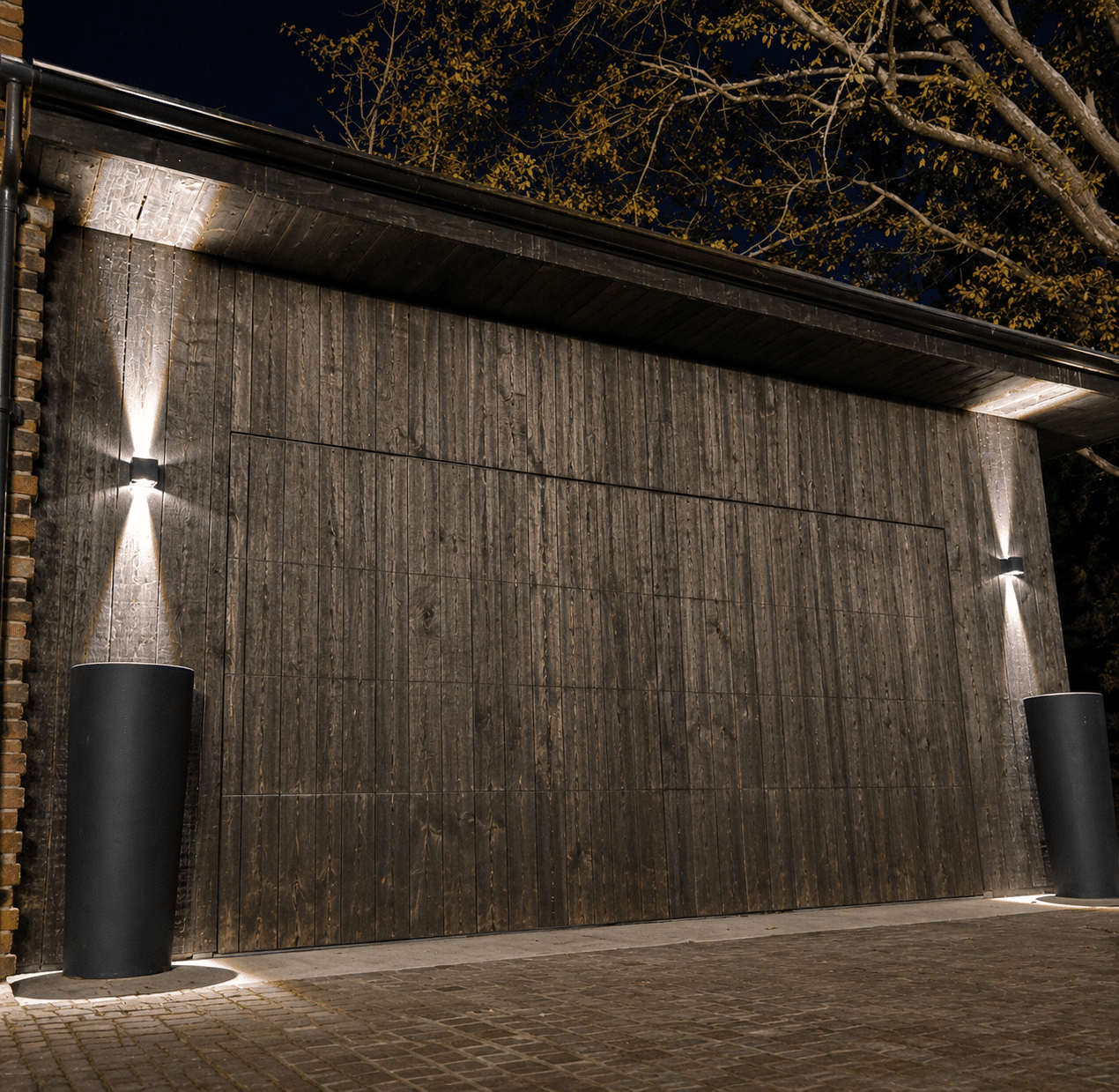 Porte de garage en bois foncé avec appliques murales lumineuses et grands pots noirs sur pavés.