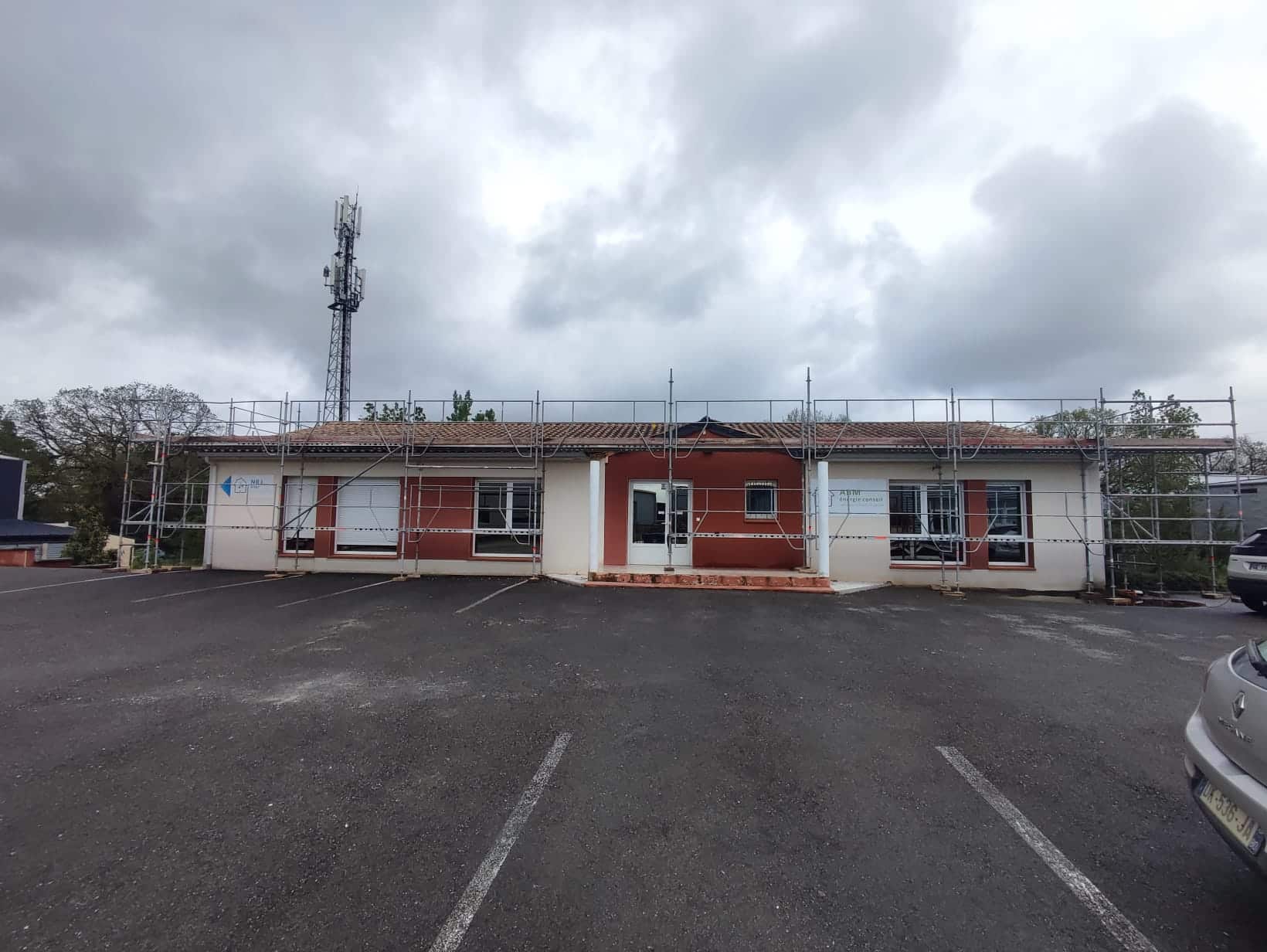 Façade ABM Énergie Conseil sous échafaudage de chantier avec parking et tour de télécommunication.