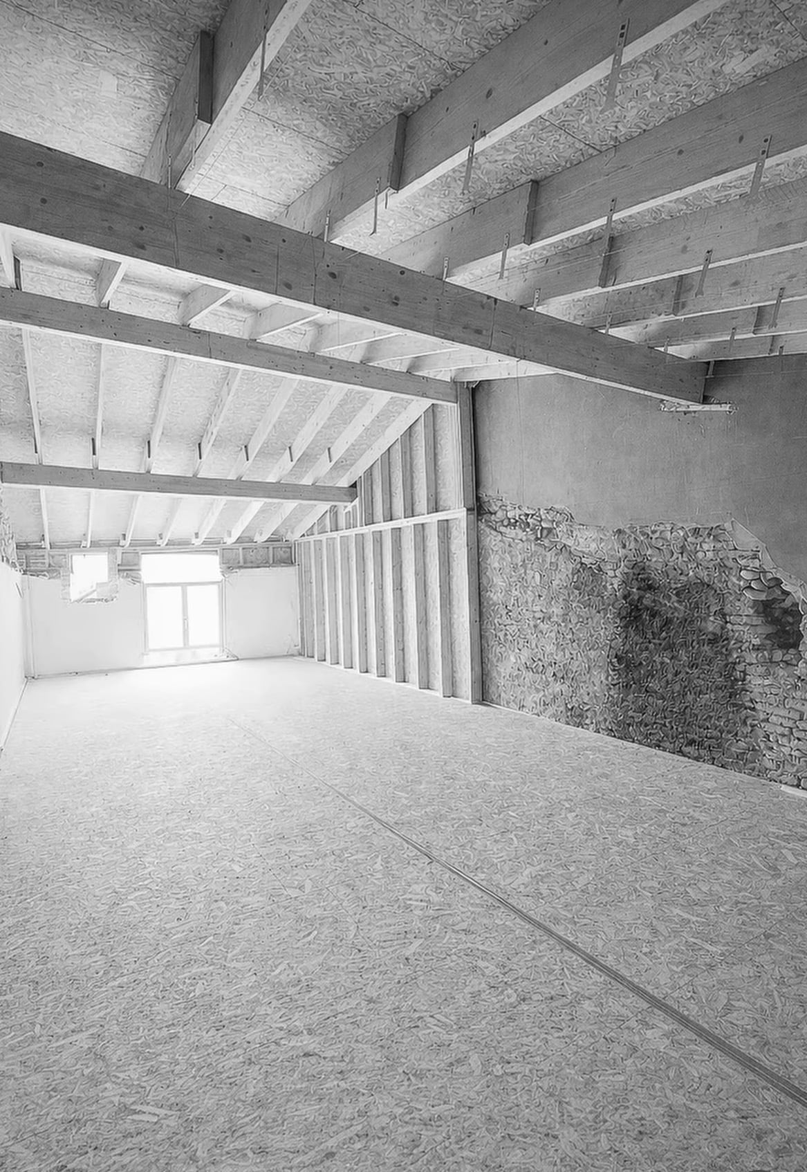 Intérieur en chantier avec poutres apparentes, sol en OSB et mur de pierre. Espace ouvert et lumineux.