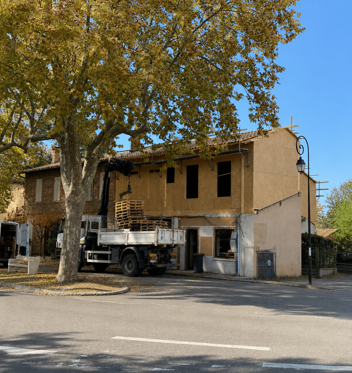 Camion grue déchargeant des palettes devant un chantier de rénovation de maison en bois sous un grand arbre.