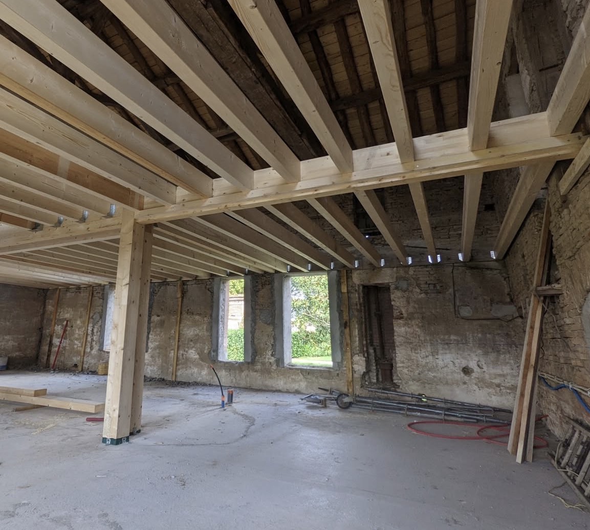 Charpente bois neuve dans bâtiment ancien en pierre, sol béton et fenêtres ouvertes.