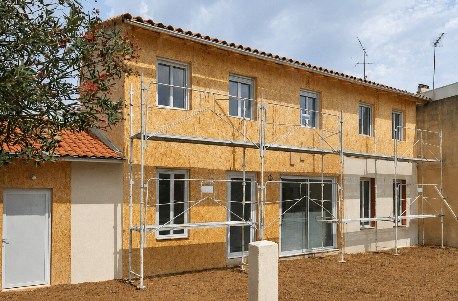 Maison en rénovation avec façade en panneaux OSB et échafaudage métallique sous un ciel nuageux.