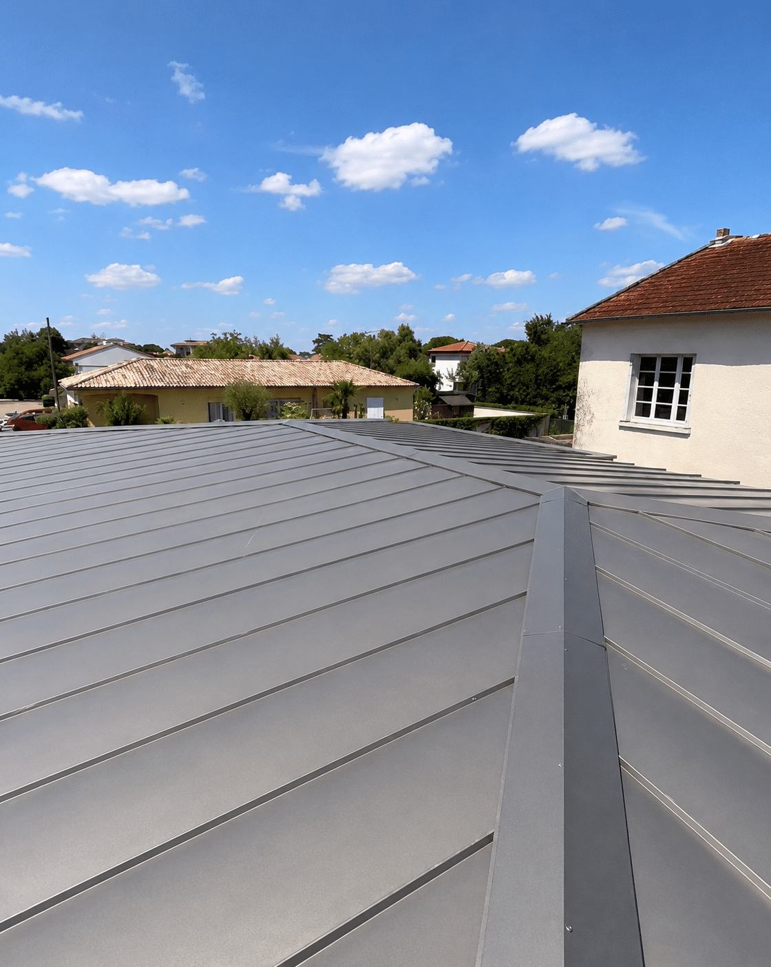 Toiture métallique grise à joint debout sous ciel bleu nuageux avec maisons en arrière-plan.