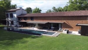 Ferme rénovée en briques, piscine miroir et terrasse moderne sur vaste pelouse.