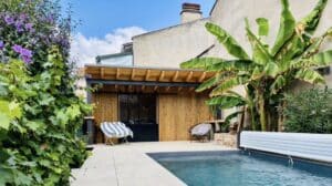 Piscine moderne avec pool house en bois, bananier et fleurs mauves dans un jardin ensoleillé.